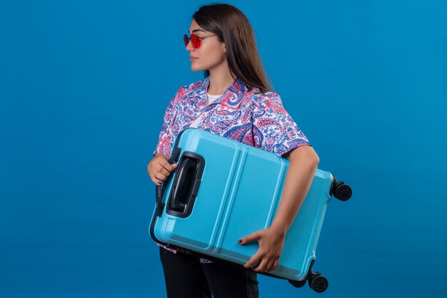 Mulher linda turista usando óculos escuros vermelhos segurando uma mala de viagem, parecendo confiante em pé de lado no espaço azul