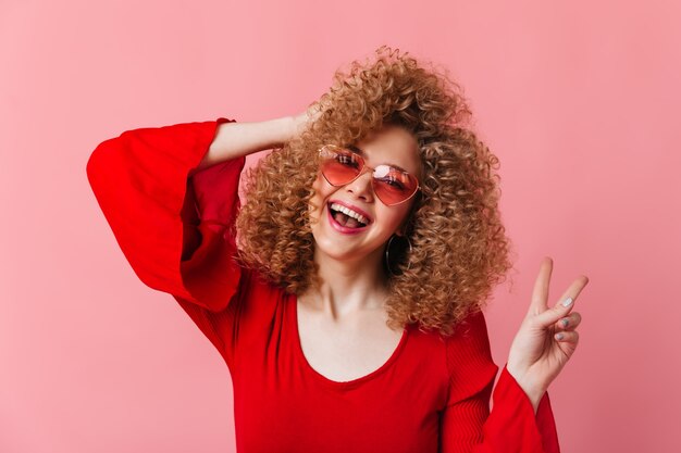 Mulher loira alegre com cachos ri e mostra o símbolo da paz no espaço rosa. Instantâneo de senhora de óculos escuros e blusa vermelha.