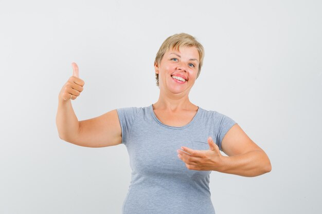 Mulher loira aparecendo os polegares e fingindo estar segurando algo na mão em uma camiseta azul clara e parecendo feliz. vista frontal.