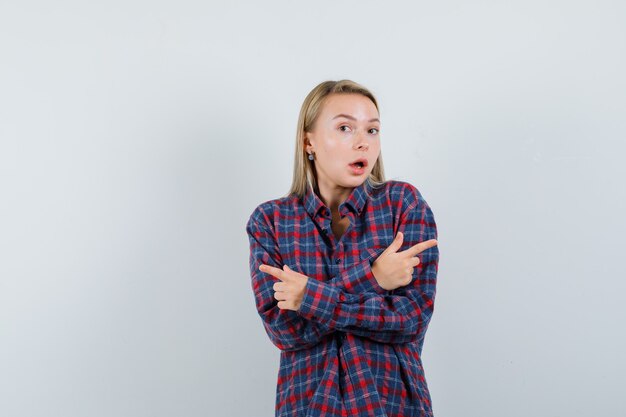 Mulher loira apontando para diferentes direções com os dois dedos indicadores em uma camisa xadrez e parecendo surpresa. vista frontal.