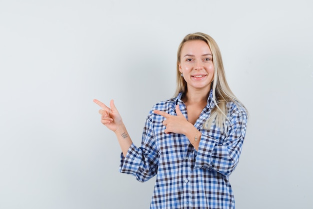 Mulher loira apontando para o canto superior esquerdo com uma camisa casual e parecendo alegre