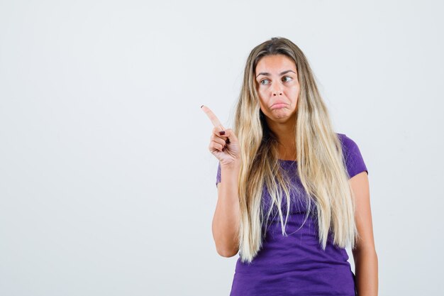 mulher loira apontando para o canto superior esquerdo de camiseta violeta e parecendo confusa, vista frontal.