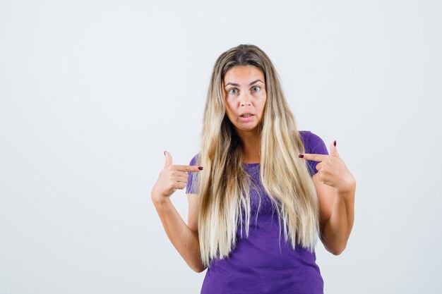 mulher loira apontando para si mesma em uma camiseta violeta e parecendo confusa, vista frontal.