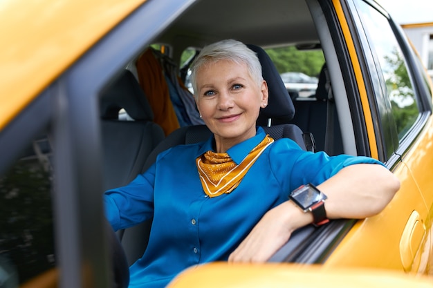 Mulher loira aposentada atraente e bem-sucedida, vestindo camisa azul e relógio de pulso, sentada confortavelmente em seu novo carro amarelo, apoiando o cotovelo na janela aberta, com uma expressão facial feliz e confiante