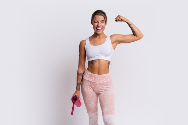 Mulher loira apta com um sorriso perfeito em uma roupa esportiva elegante, olhando para a câmera e segurando uma garrafa de água sobre a parede branca. Demonstre músculos.