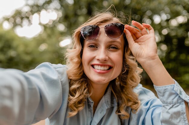 Mulher loira atraente e sorridente andando no parque com roupa de verão tirando foto de selfie no telefone