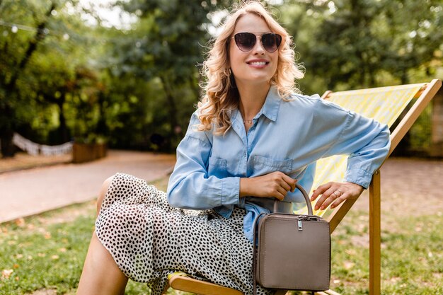 Mulher loira atraente e sorridente sentada em uma espreguiçadeira com roupa elegante