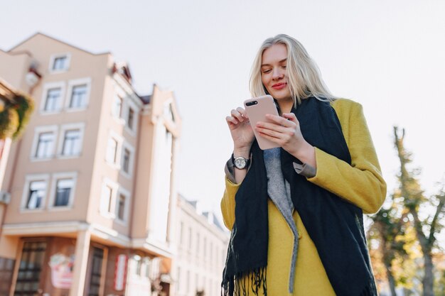 Mulher loira atraente emocional fofa com casaco e smartphone caminhando pela cidade
