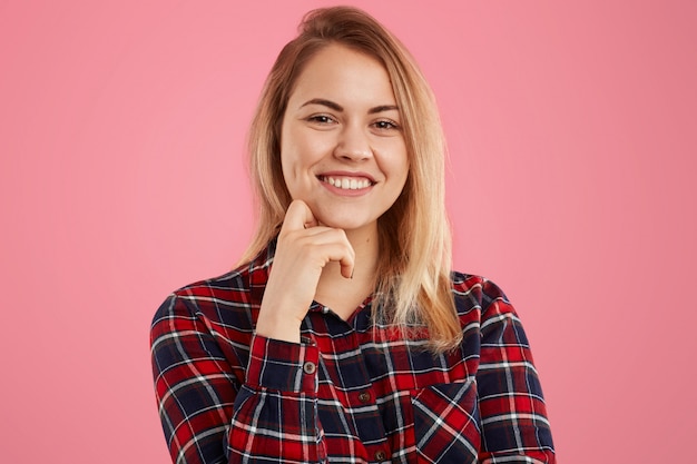 Mulher loira bonita com sorriso gentil, mantém uma mão sob o queixo, vestida com camisa quadriculada