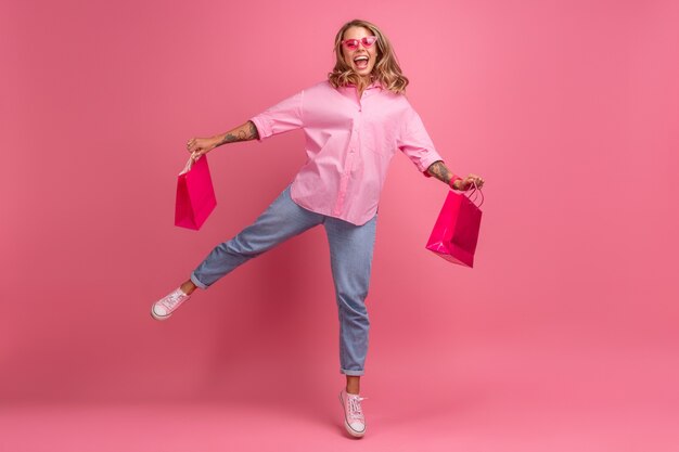 Mulher loira bonita em uma camisa rosa e calça jeans sorrindo pulando no fundo rosa isolada se divertindo segurando sacolas de compras