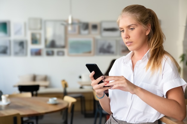 Foto grátis mulher loira bonita focada vestindo camisa branca, usando smartphone em pé no espaço de trabalho