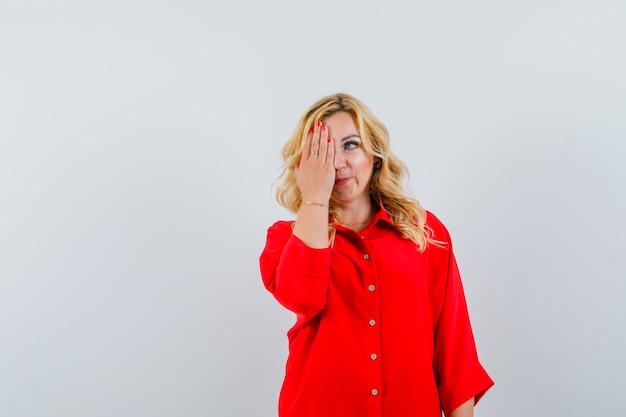 Mulher loira cobrindo os olhos com uma mão na blusa vermelha e parecendo feliz.