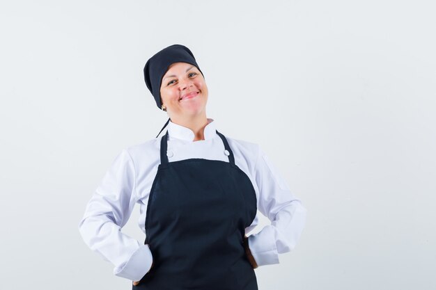 Mulher loira colocando as mãos na cintura em uniforme preto de cozinheiro e bonita, vista frontal.