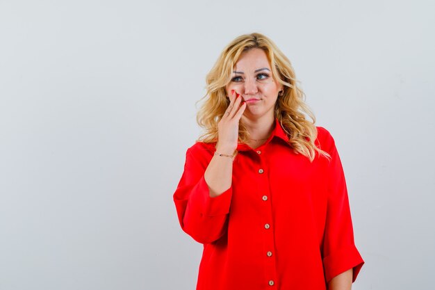 Mulher loira com blusa vermelha colocando a mão na bochecha, olhando para longe e pensativa