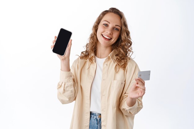 Mulher loira com cabelo encaracolado, mostra tela vazia do smartphone e cartão de crédito, compra na internet, demonstra um app, parede branca