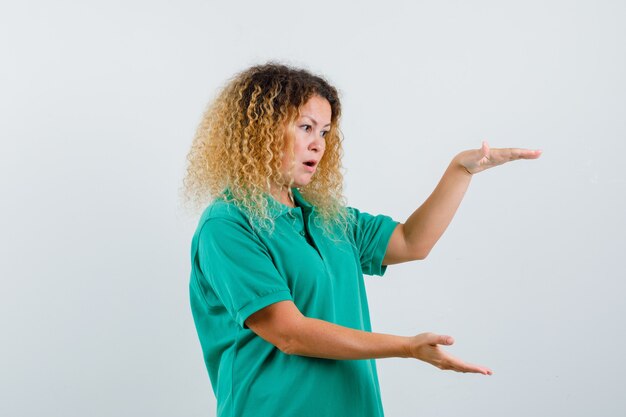 Mulher loira com cabelo encaracolado, mostrando sinal de tamanho grande em uma camiseta verde e parecendo surpresa, vista frontal.