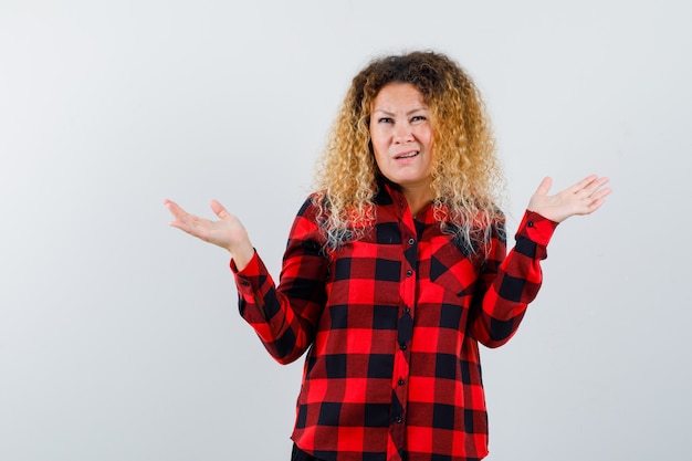 Mulher loira com cabelo encaracolado, mostrando um gesto desamparado em camisa xadrez e parecendo confusa, vista frontal.