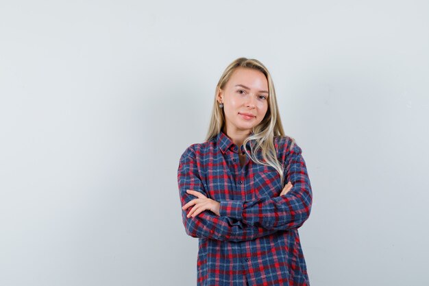 Mulher loira com camisa xadrez, segurando os braços cruzados e olhando atraente, vista frontal.