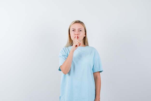 Mulher loira com camiseta azul mostrando gesto de silêncio e parecendo séria