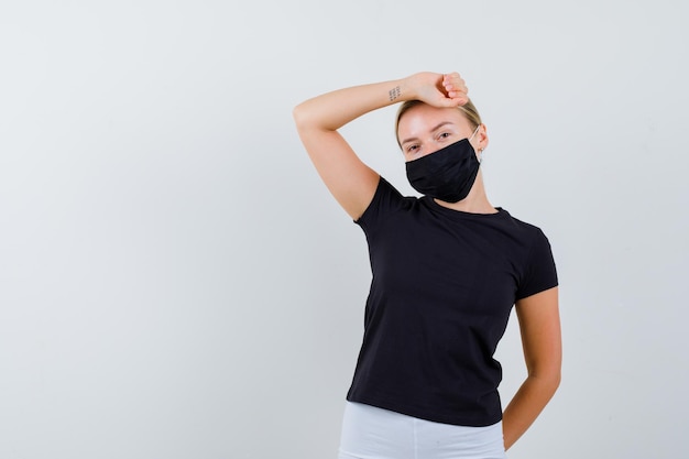 Mulher loira com camiseta preta, calça branca, máscara preta levantando o braço acima da cabeça