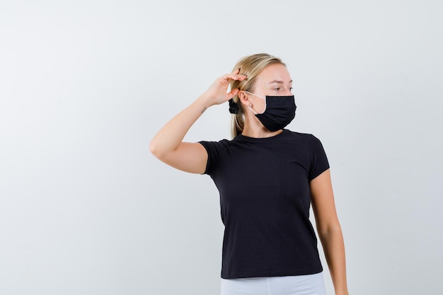 Mulher loira com camiseta preta, calça branca, máscara preta segurando a mão perto da orelha