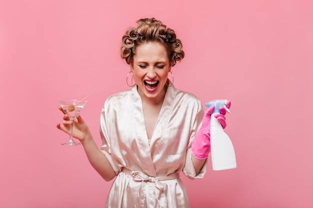 Mulher loira com um robe rosa posando com um copo de martini e detergente