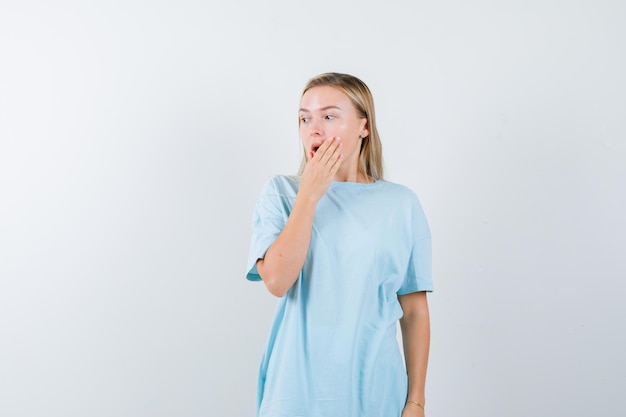 Mulher loira com uma camiseta azul cobrindo a boca com a mão, olhando para longe e parecendo surpresa