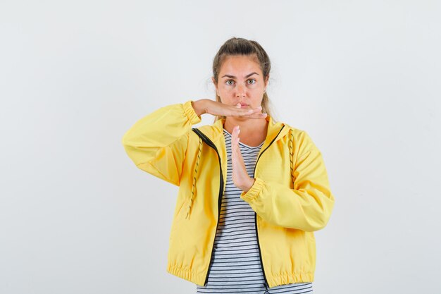 Mulher loira com uma jaqueta militar amarela e uma camisa listrada mostrando um gesto de pausa e parecendo séria