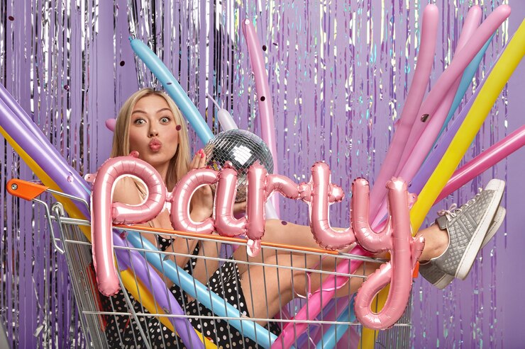 Foto grátis mulher loira de aparência agradável e engraçada no carrinho de compras com balões de modelagem longos e bola de discoteca, usa vestido e sapatos esportivos, olha para a câmera com os lábios dobrados, se diverte em uma festa barulhenta com os colegas