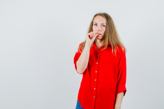 Mulher loira de camisa vermelha roendo a unha e parecendo pensativa,