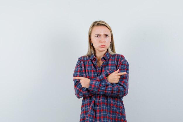 Mulher loira de camisa xadrez, apontando para diferentes direções com o dedo indicador e olhando furiosa, vista frontal.