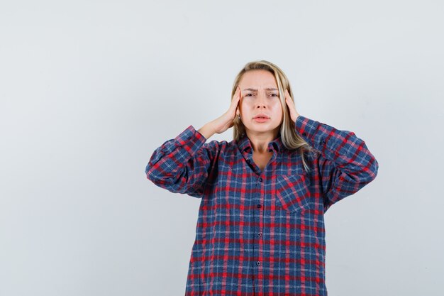 Mulher loira de camisa xadrez, de mãos dadas na cabeça e parecendo exausta, vista frontal.