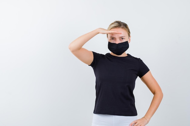 Mulher loira de camiseta preta, calça branca, máscara preta olhando para longe