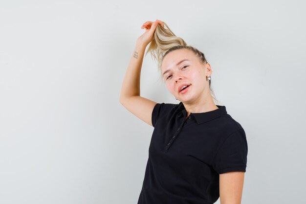 Mulher loira de camiseta preta segurando o cabelo e parecendo feliz