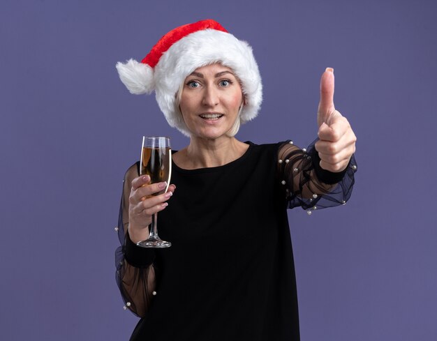 Mulher loira de meia-idade alegre com chapéu de Natal, olhando para a câmera, segurando a taça de champanhe, mostrando o polegar isolado no fundo roxo