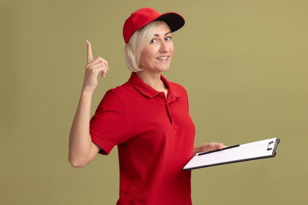 Mulher loira de meia-idade, alegre, entregadora de uniforme vermelho e boné em pé, segurando uma prancheta e um lápis apontando para cima