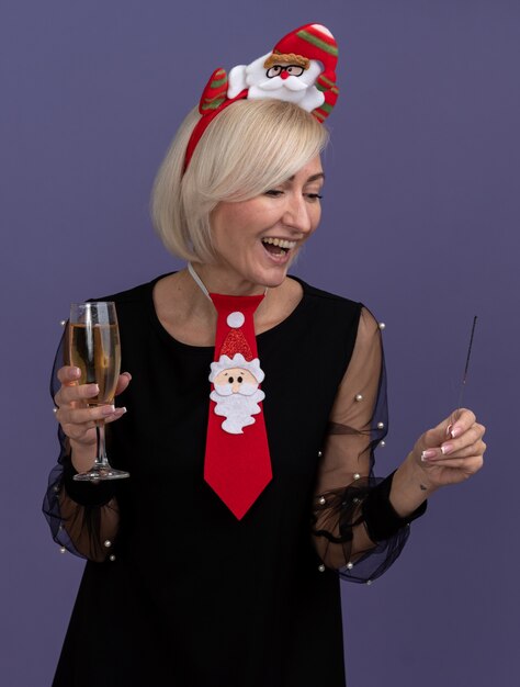 Foto grátis mulher loira de meia-idade alegre usando bandana de papai noel e gravata segurando um diamante de feriado e uma taça de champanhe, olhando para um diamante isolado na parede roxa