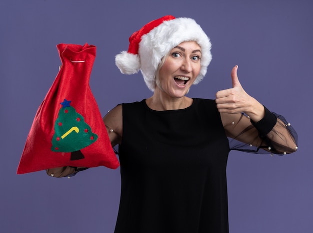 Mulher loira de meia-idade impressionada com chapéu de Natal segurando um saco de Natal olhando para a câmera mostrando o polegar isolado no fundo roxo