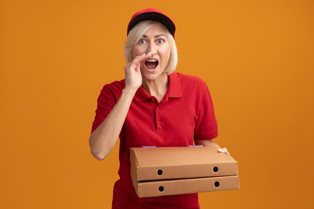 Mulher loira de meia-idade impressionada, entregadora de uniforme vermelho e boné, segurando pacotes de pizza, mantendo a mão perto da boca e sussurrando