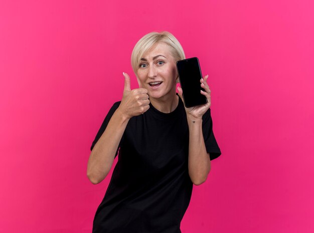 Mulher loira de meia-idade impressionada mostrando o celular e o polegar para cima, olhando para a frente, isolada na parede rosa
