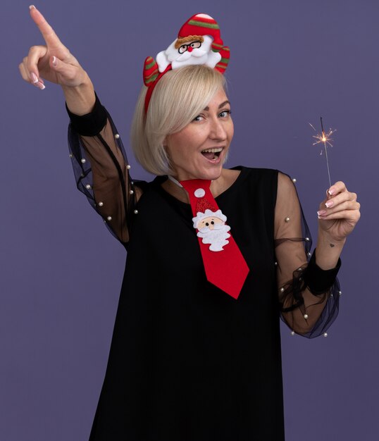 Mulher loira de meia-idade impressionada usando bandana de papai noel e gravata segurando um diamante de feriado apontando para cima, isolado na parede roxa