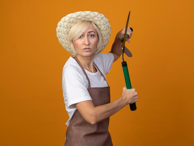 Mulher loira de meia-idade, jardineira, impressionada, de uniforme, usando um chapéu em pé na vista de perfil, segurando uma tesoura de cerca viva isolada na parede laranja com espaço de cópia