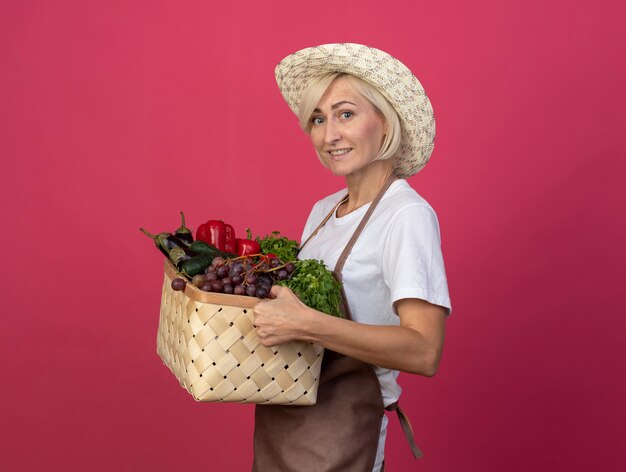 Mulher loira de meia-idade, jardineira, sorridente, de uniforme, usando um chapéu em pé na vista de perfil, segurando uma cesta de legumes, olhando para a frente, isolada na parede vermelha