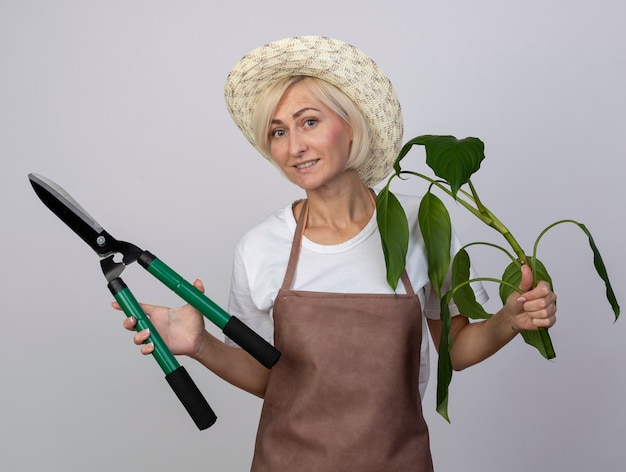 Mulher loira de meia-idade, jardineira sorridente, de uniforme, usando um chapéu, segurando uma tesoura de planta e uma tesoura de sebe isolada na parede branca