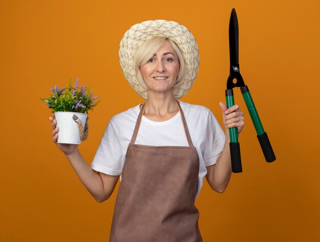 Mulher loira de meia-idade, jardineira sorridente, de uniforme, usando um chapéu, segurando uma tesoura e um vaso de flores, olhando para a frente, isolada em uma parede laranja