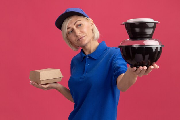 Mulher loira de meia-idade satisfeita, entregadora de uniforme azul e boné, segurando um pacote de comida esticando os recipientes de comida para a frente, olhando para eles isolados na parede rosa