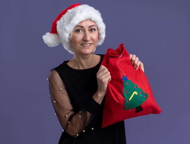 Mulher loira de meia-idade sorridente com chapéu de Natal segurando um saco de Natal olhando para a câmera isolada no fundo roxo