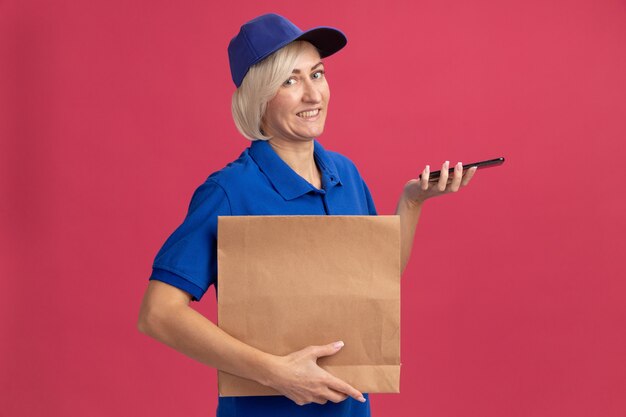 Mulher loira de meia-idade sorridente, entregadora de uniforme azul e boné, segurando um pacote de papel e um telefone celular