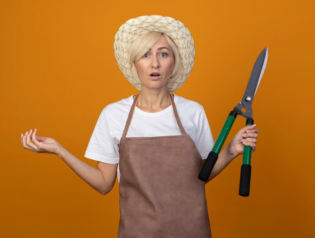 Foto grátis mulher loira de meia-idade, surpreendida, de uniforme, usando um chapéu, segurando uma tesoura, mostrando a mão vazia, isolada na parede laranja