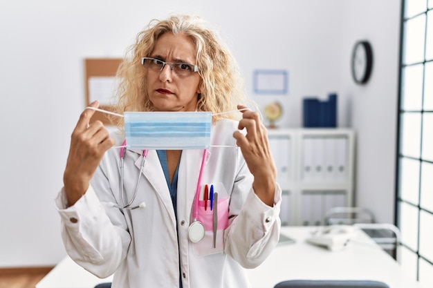 Mulher loira de meia-idade vestindo uniforme médico e máscara médica cética e nervosa, franzindo a testa chateada por causa do problema. pessoa negativa.